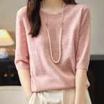 A woman wearing a pink crewneck merino wool sweater with half sleeves, paired with white trousers and accessorized with a long pearl necklace.