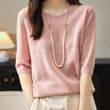 A woman wearing a pink crewneck merino wool sweater with half sleeves, paired with white trousers and accessorized with a long pearl necklace.