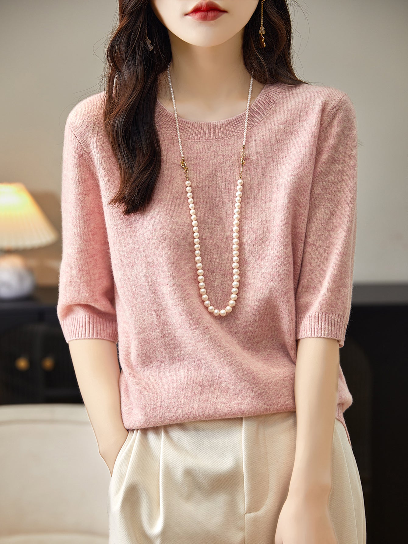 A woman wearing a pink crewneck merino wool sweater with half sleeves, paired with white trousers and accessorized with a long pearl necklace.