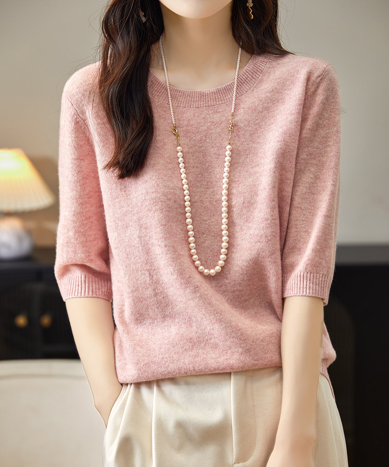 A woman wearing a pink crewneck merino wool sweater with half sleeves, paired with white trousers and accessorized with a long pearl necklace.