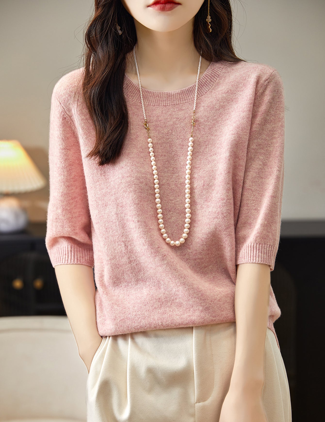 A woman wearing a pink crewneck merino wool sweater with half sleeves, paired with white trousers and accessorized with a long pearl necklace.